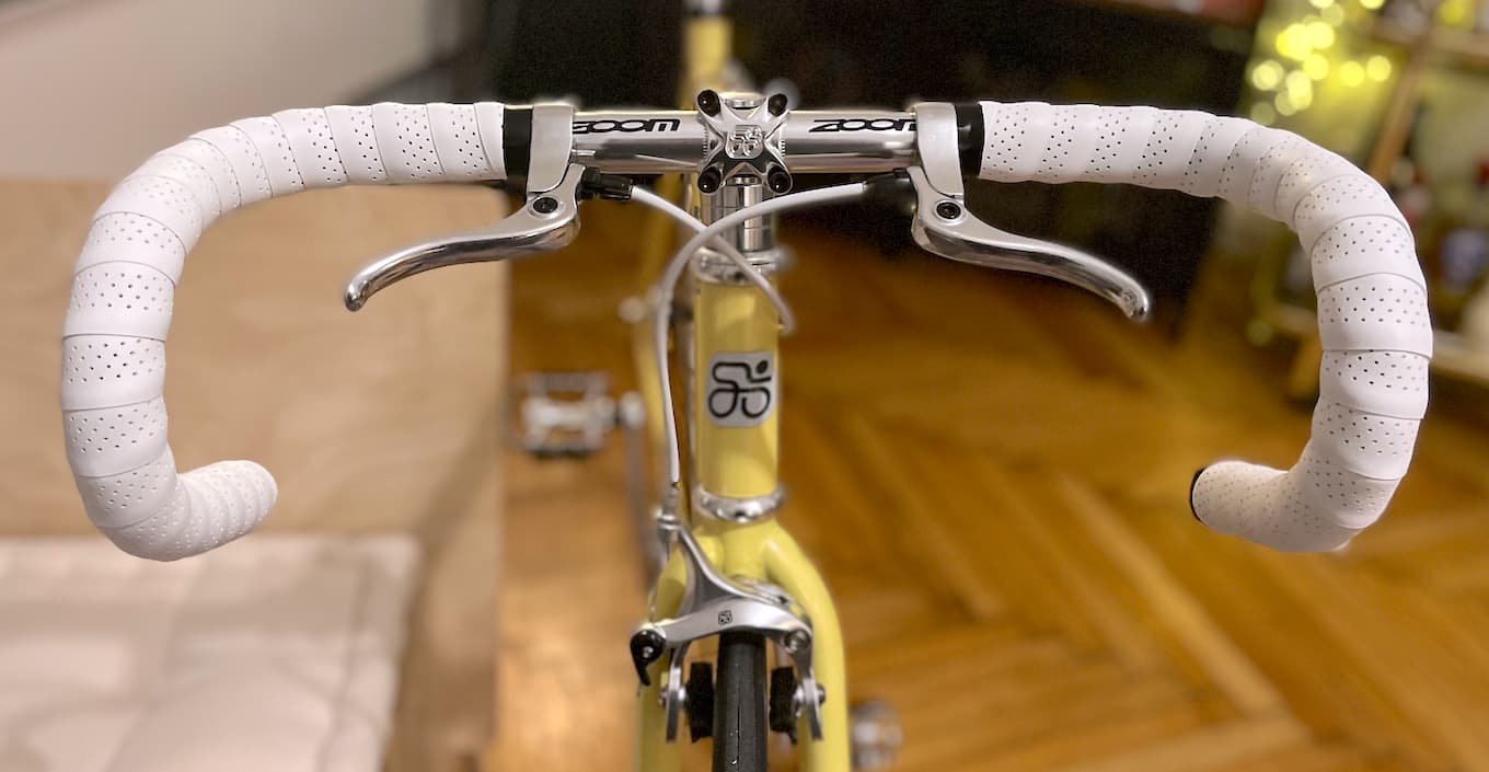 A bicycle cockpit from the front: drop bars with white perforated bar tape, brake levers and white cabling.